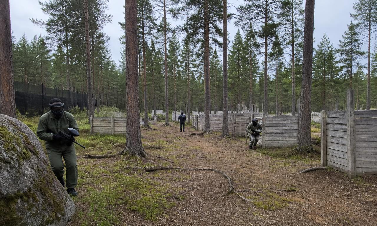 Laaja näkymä metsäiselle paintball-kentälle, jossa pelaajia puisten suojien välissä.